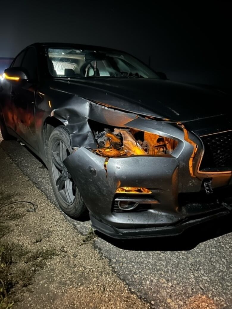 A dark grey car sits on a road. The front of the car is broken and dented.