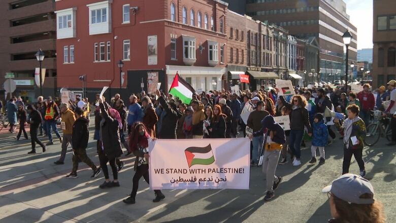 A crowd marches behind a large white sign with black letters reading 