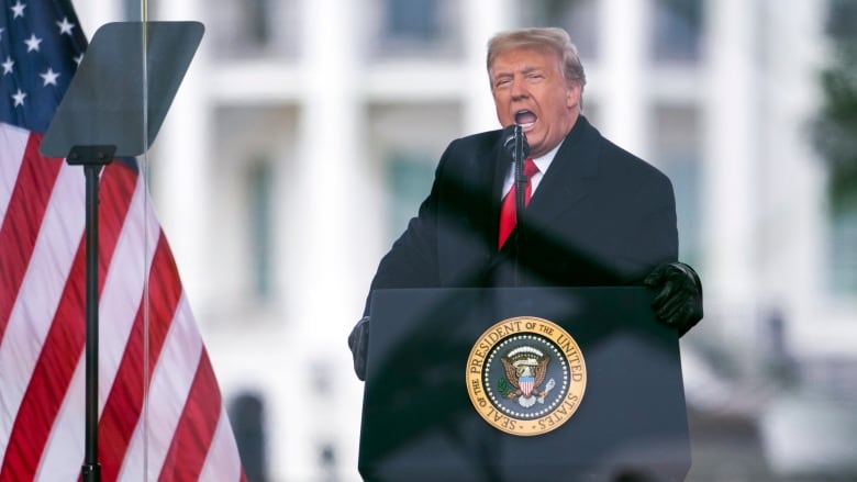 A man in a long winter coat wearing a tie speaks at a podium bearing the seal, 'The President of the United States of America.' He is behind a glass partition and in front of an American flag.