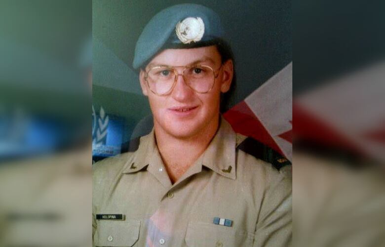 A man in a beret and army shirt wears glasses and smiles at the camera. He is seen from the mid-chest to the top of the head.