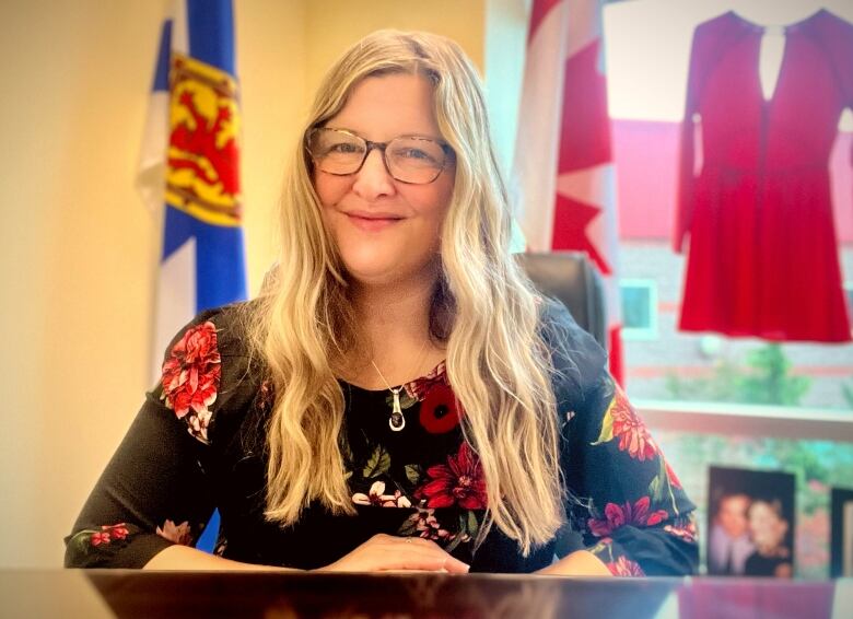 A woman with blonde hair and glasses and a dark flower-print dress sits at a desk in front of Canada and Nova Scotia flags.