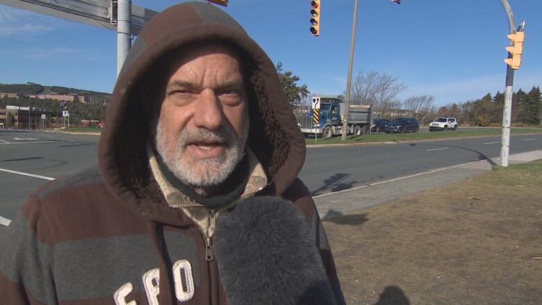 A man wearing a brown hoodie with the hood pulled up stands on the sidewalk at an intersection.