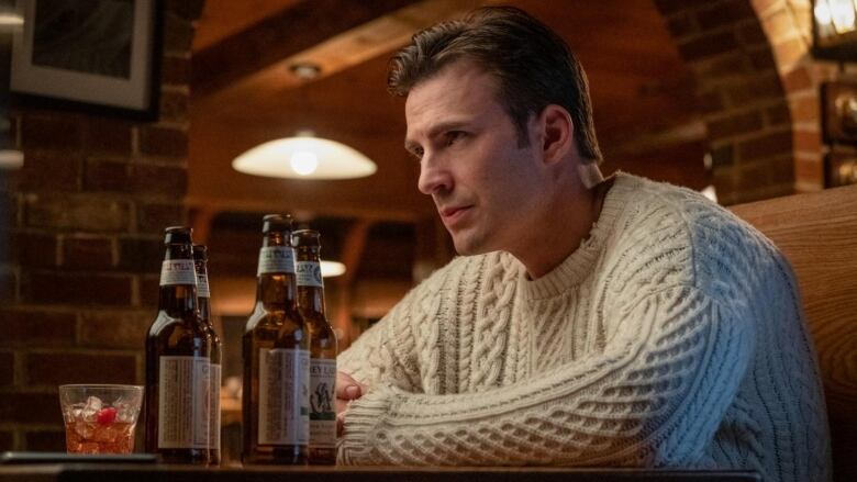 Cloesup on Chris Evans in the film Knives Out. He's wearing an off-white knitted sweater and sitting at a table with beer bottles on it in a pub. 