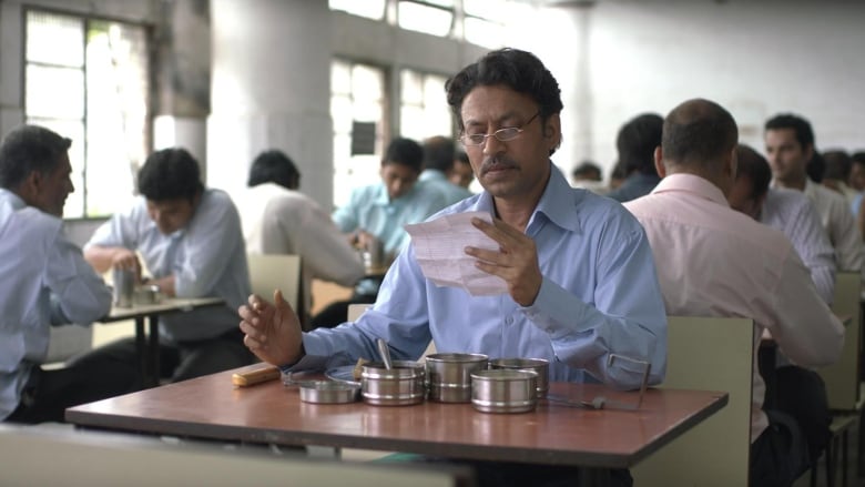 Irrfan Khan in the film The Lunchbox. He's wearing a blue shirt, sitting by himself at a table in a lunchroom, reading a note on a piece of paper, with a silver lunchbox in front of him.   