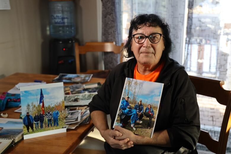 A woman with dark brown hair and glasses, wearing a black hoodie and orange t-shirt, sits at her kitchen table holding a family photograph.