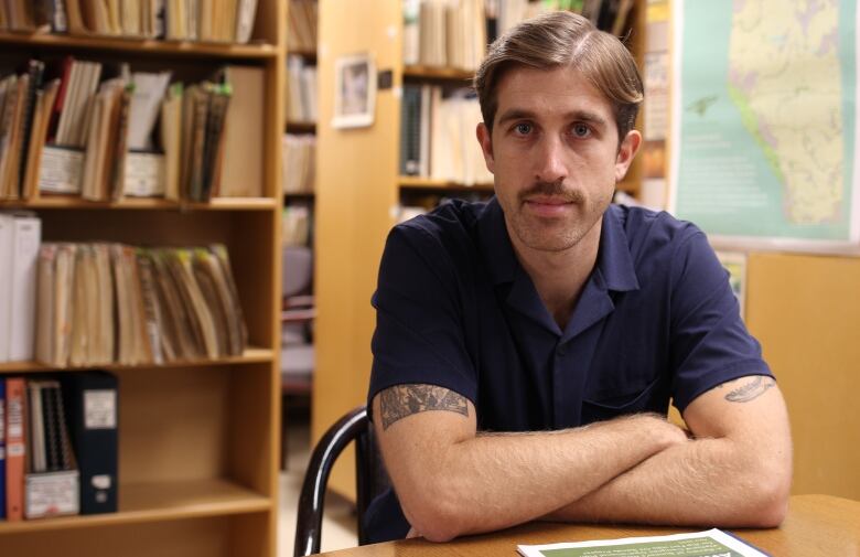 A man with a moustache and a blue button-down shirt is pictured with his arms folded inside a library.