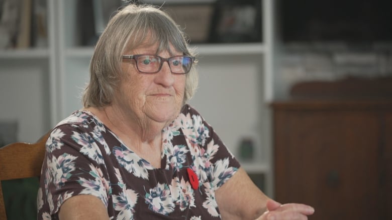 A woman with grey shoulder-length hair and glasses, wears a floral-patterned dress with a poppie pinned to it.