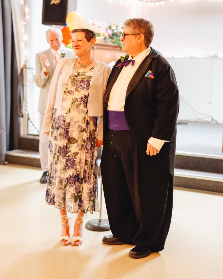 A beaming woman in a formal dress stands hand in hand with another woman dressed in a tuxedo and rainbow pocket square. In the background, a man in a suit and boutonniere smiles and claps.