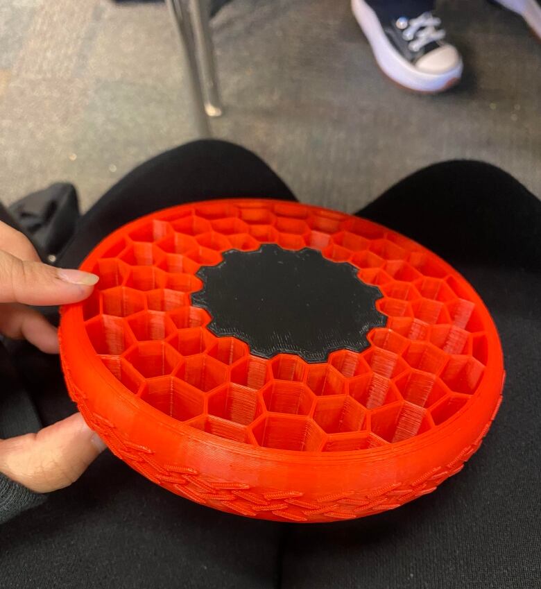 A red plastic wheel about the size of a shopping cart wheel sits on the lap of a teenager wearing black pants.