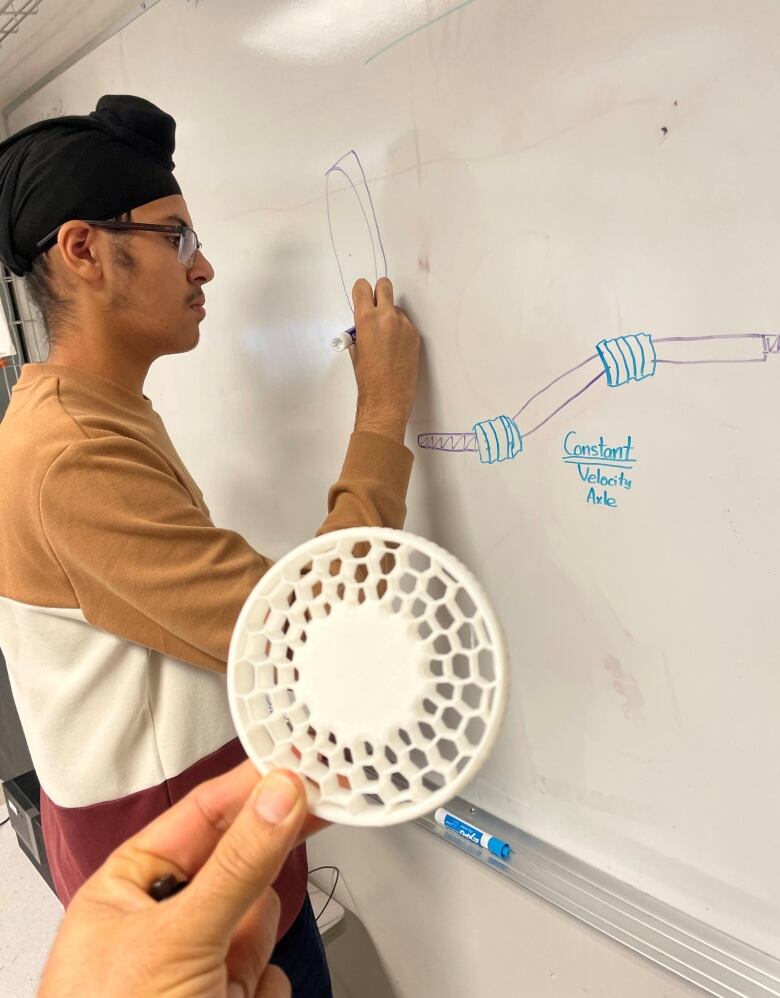 A South Asian teenage boy stands at a white board drawing wheel axles. Next to him, a student's hand holds up a 3-D printed red plastic wheel.