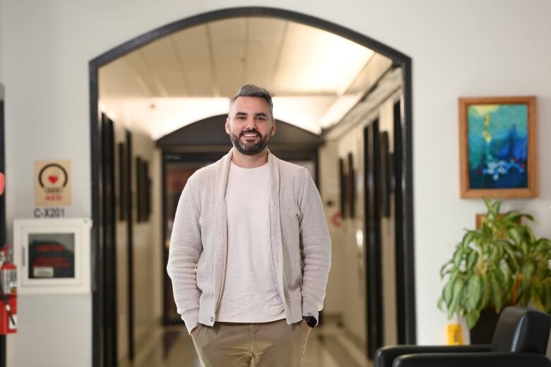 A man with with salt and pepper hair and a beard stands in a lit hallway, wearing a white t-shirt and beige cardigan. He is smiling.