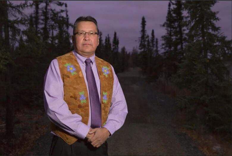 A man standing in a forest with his hands together in front of him. He is wearing a suede vest with blue flowers and glasses.