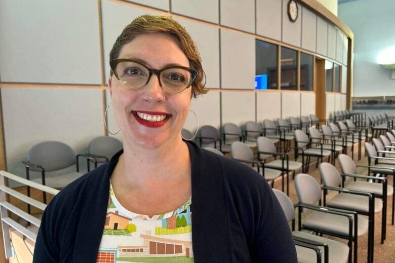 A woman with short red hair and large glasses stands in front of rows of empty chairs