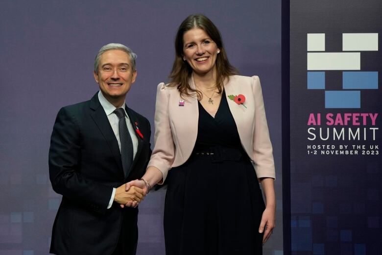 A man in a navy suit and dark tie shakes hands with a woman in a light pink blazer.