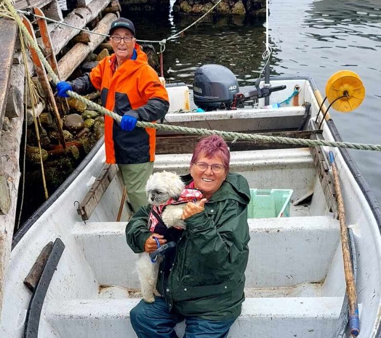 A woman sits in the middle of a small dory. She is holding a small, fluffy dog. A fisherman stands behind them, untying the boat from a wharf.