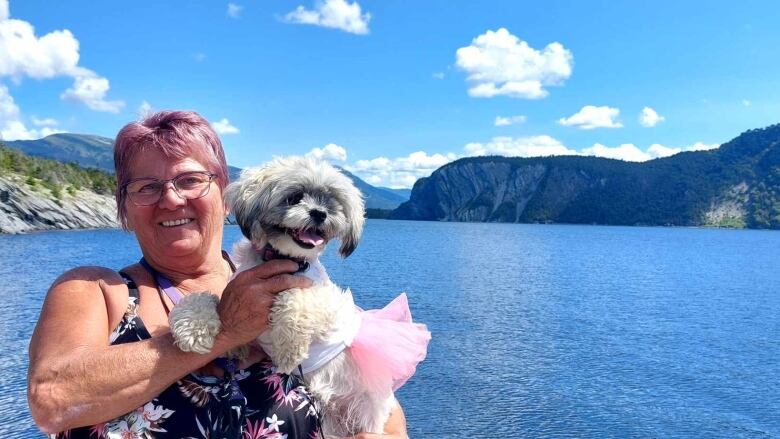 A photo of a smiling woman holding a dog. She is standing near the ocean. There are mountains and hills in the background. 