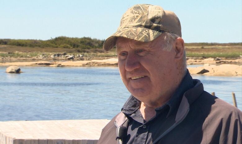 Close up head shot of Paul Bromley in the foreground. He's wearing a hat. A wharf and the seaside is in the background. 