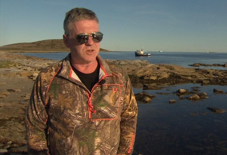 Wayne Barney, wearing sunglasses and camouflage clothing, stands on the rocky seaside. A small fishing boat is in the background.  