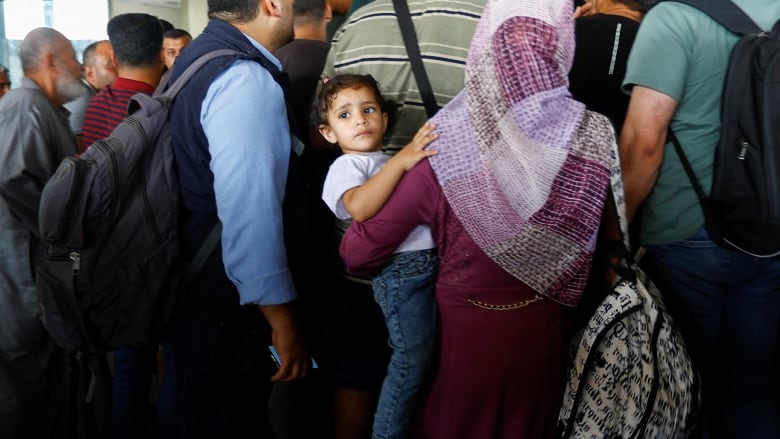 A woman holds a toddler on her hip as they wait in a crowded room at a border crossing.