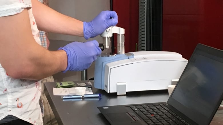 A technician wearing blue rubber gloves is using what looks like a microscope to check samples of drugs users provide. A laptop displays the results.