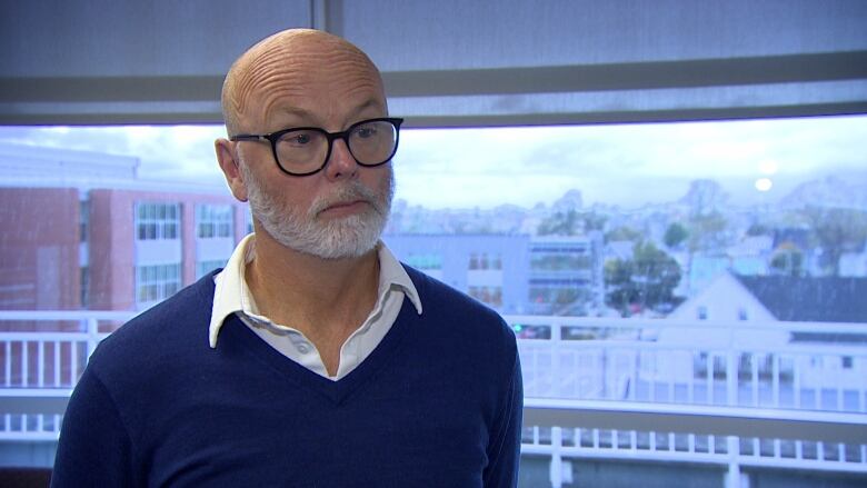 A bald man with a white beard and glasses wears a purper sweater overtop a white, collard shirt. He stands in front of a window with a cityscape in the background. 