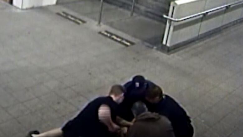 An image taken from grainy CCTV footage shows four police officers crowded in the bottom right corner, holding someone on the ground. One of them can be seen winding up his right arm for a punch. 