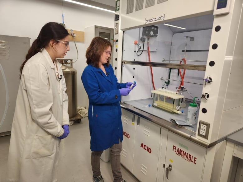 Two women in lab coats handle a sample beside a machine.