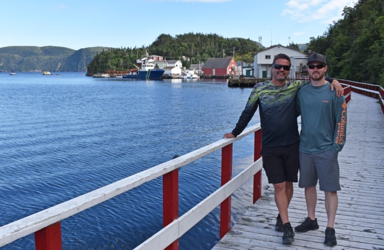 Two men stand on a wooden boardwalk with a red and white railing beside a body of water. Boats and houses can be seen in the background.