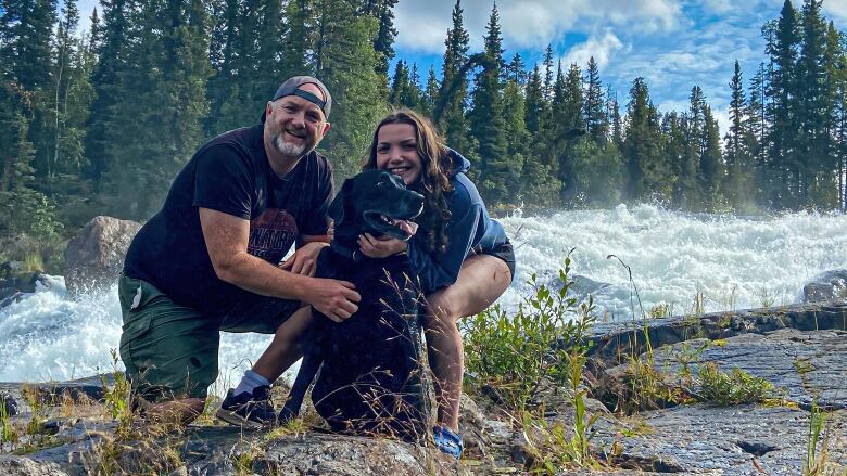 Two people pose with a dog in front of rapids.