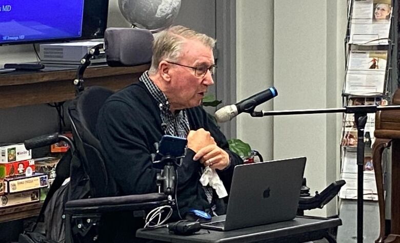 Man with grey hair and glasses wearing checked black and white shirt and black cardigan sits in wheelchair, speaking into microphone with Apple laptop on desk in front of him.