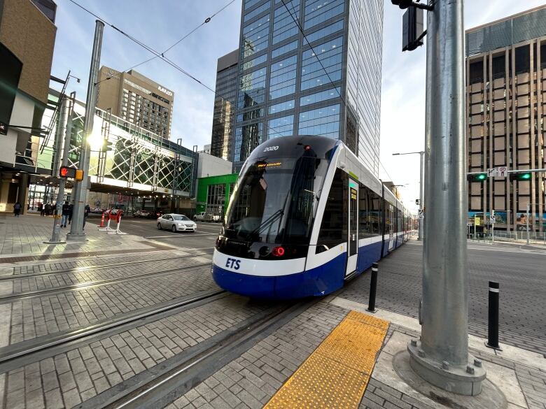 A blue and white train rolls across some tracks. Various buildings are in the background.