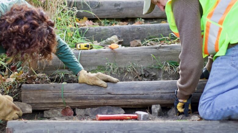 Two people hold a plank of wood in place on steps in a hill while one pushes rocks under the front of it. 