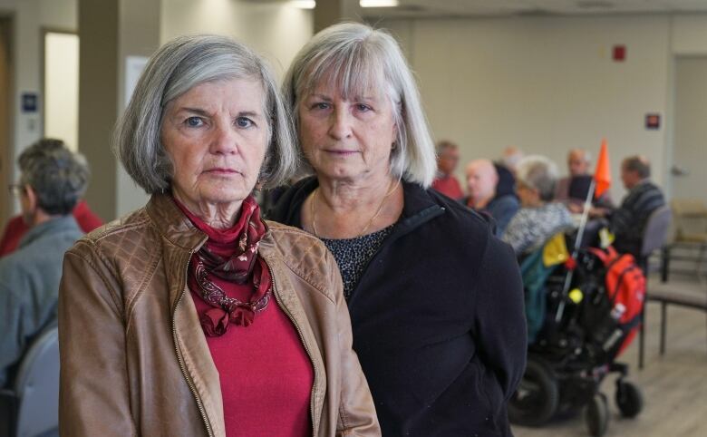 Janet White, at left, and Christine Heron, right, are coordinators of a bridge group that plays twice a week at the Heron Road Community Centre. They stand in their meeting room as about two dozen players, mostly seniors, sit at tables behind them.