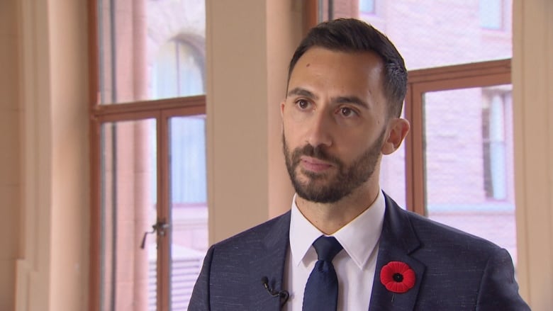 A man in a suit and tie, with a poppy on his lapel, looks left off-camera during an interview in an interior hallway of a historic building.