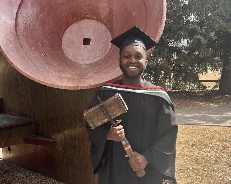 A smiling young man wearing a graduates cap and gown holds a wooden mallet while standing in front of a large, bell-shaped sculpture.