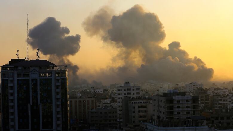 Two large smoke clouds rise into the yellow sky above the concrete buildings of a city.