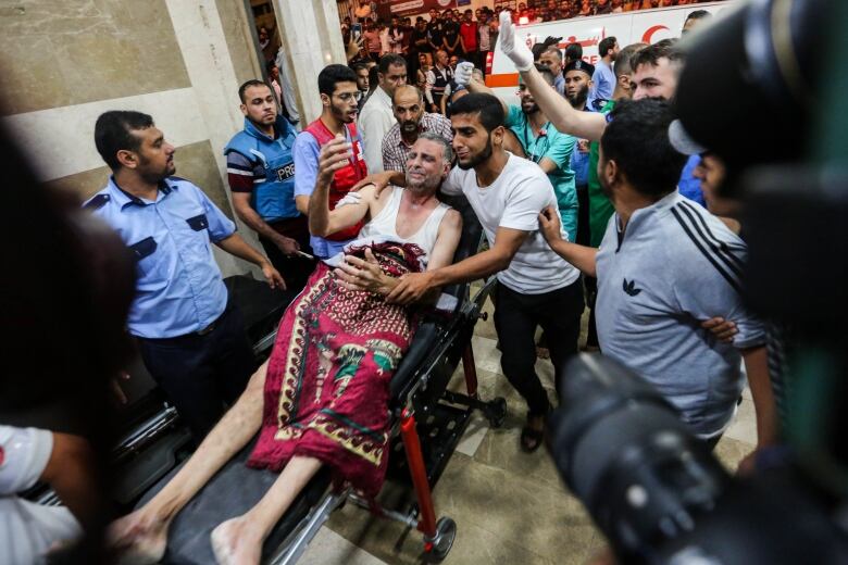 A man covered in a blanket lays on a stretcher with several people pushing him inside a hospital. A crowd of people and an ambulance are in the background.