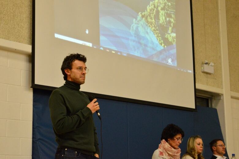 A man in a green turtleneck sweater holds a microphone. 