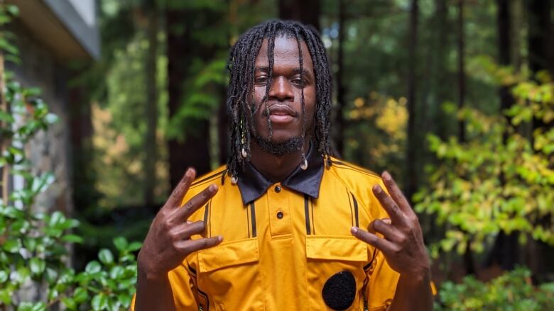 A Black man makes a peace hand gesture with both hands. He is wearing a yellow shirt and has dreads.