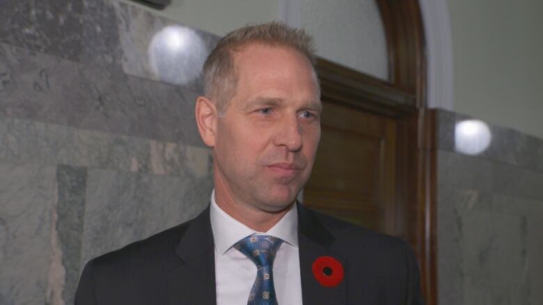 A man with blonde hair wearing a suit stands in a hallway, wearing a poppy on his jacket.