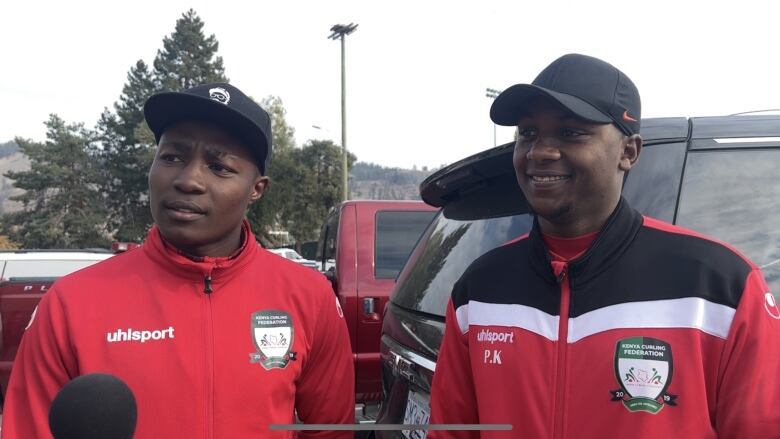 Two Black men wearing red jackets speak to a camera.