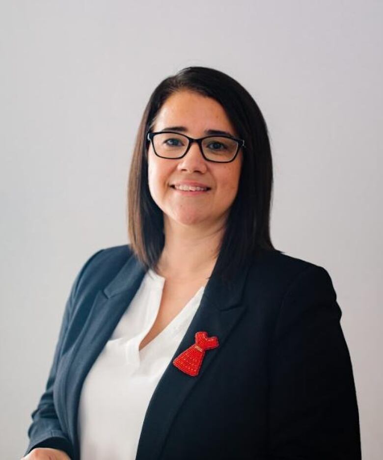 A woman wearing glasses stands for a photo. She is wearing a white blouse, dark blazer, and a beaded pin in the shape of a red dress.