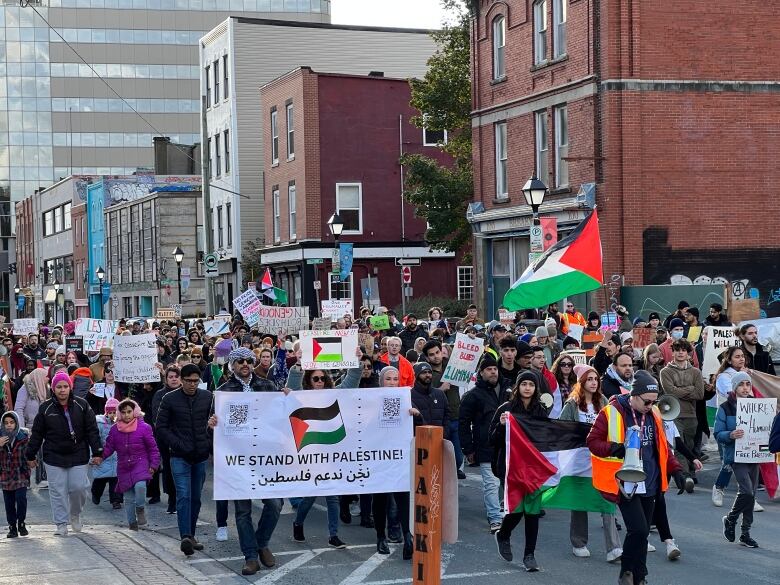 Hundreds of protesters carrying signs and flags are gathered on a downtown city street.