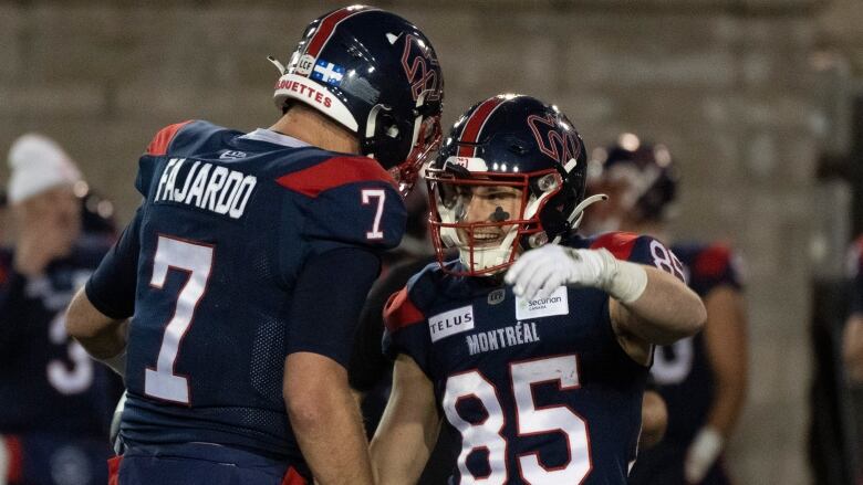 A male football receiver wraps his arms around his team's quarterback in celebration while smiling during a game.