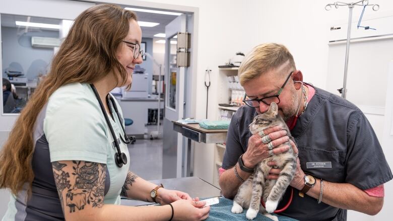 a woman in blue scr8ibs helps a  man in grey scrubs care for a short haired cat on an examination table 