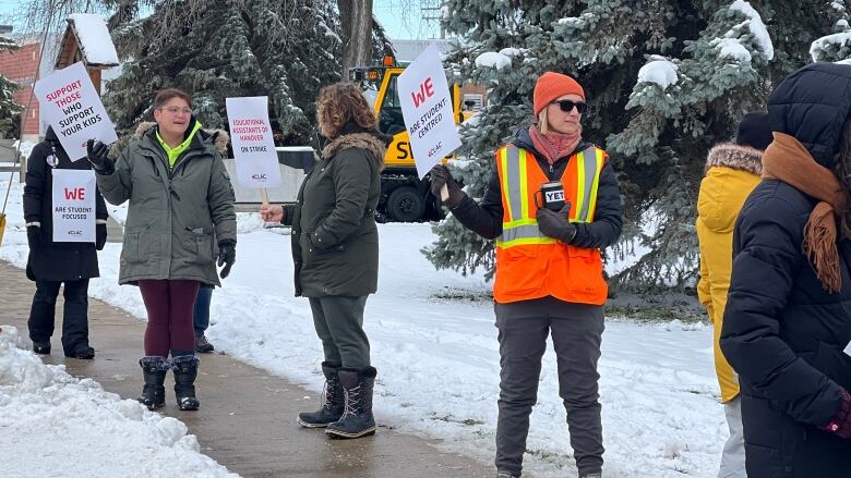 People hold picket signs and stand on the sidewalk outside a park.