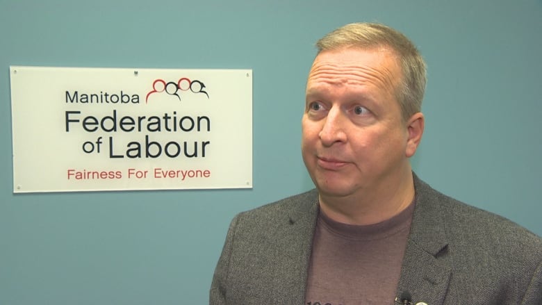 A man in a blazer stands in front of a wall with the Manitoba Federation of Labour signage on it.