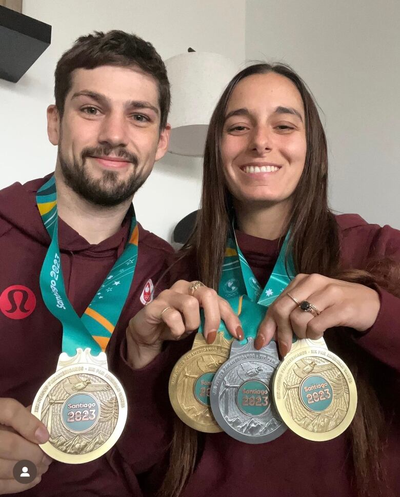 Two people wearing burgundy sweaters hold up gold, silver and bronze medals that are hanging around their necks.