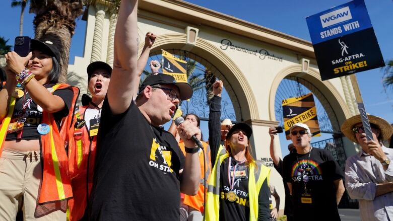 A group of chearing people wearing high-visibility vests stand outside of an arch adorned with the words 