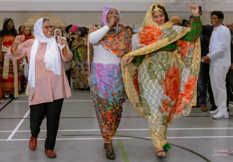 Three women wearing colourful dresses walk through a gym. 